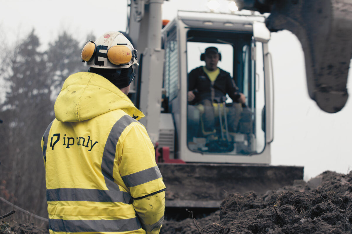 Man i varseljacka med IP-Onlys logga tittar mot en man i grävmaskin