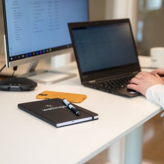 Närbild på ett par händer som arbetar med en laptop. På bordet syns också ett skrivblock med Welingos logga.